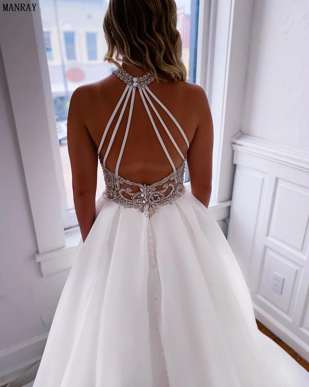Back view of a woman in a white wedding gown with a beaded halter neckline and strappy back. The bodice features intricate lace and bead detailing, with a long flowing skirt.
