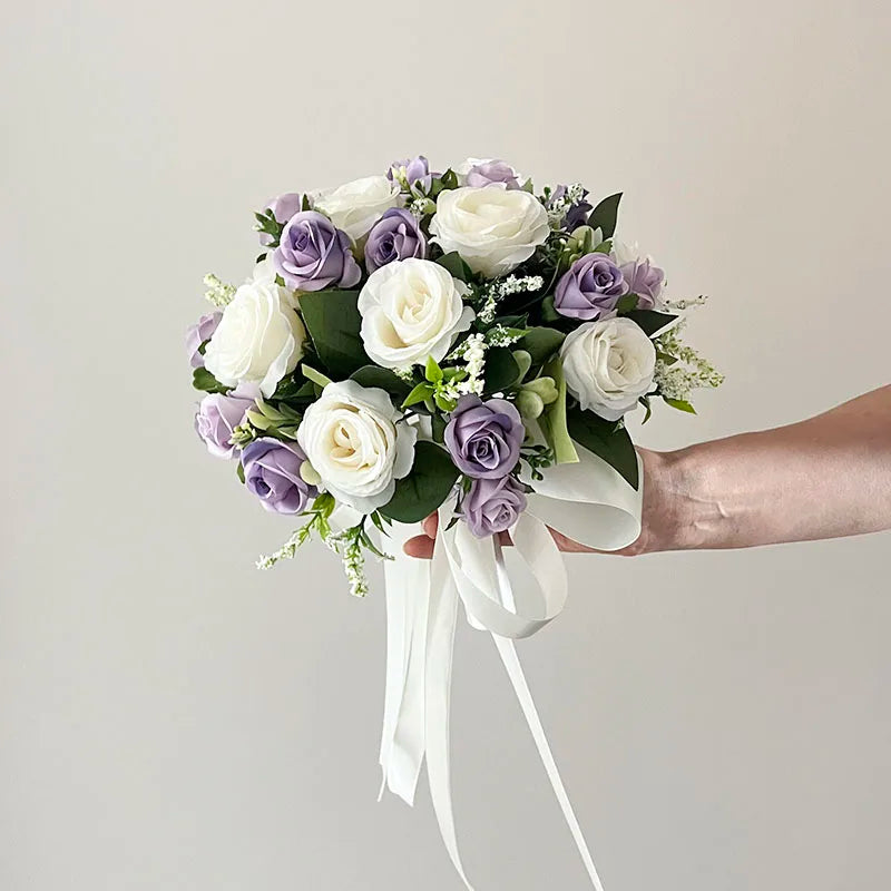 A hand holds a bouquet of white and purple roses with green leaves, adorned with white ribbons, against a plain white background.
