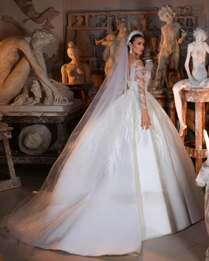 A bride in an elegant white gown with intricate lace and a long veil stands among classical sculptures in an art studio, looking over her shoulder.
