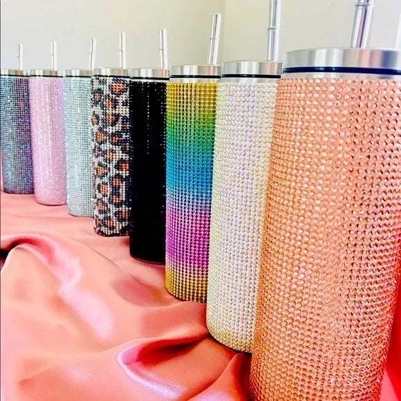 Row of stainless steel tumblers, each covered in different colored rhinestones, standing on a silky pink fabric. The colors range from pastel to bold leopard patterns.