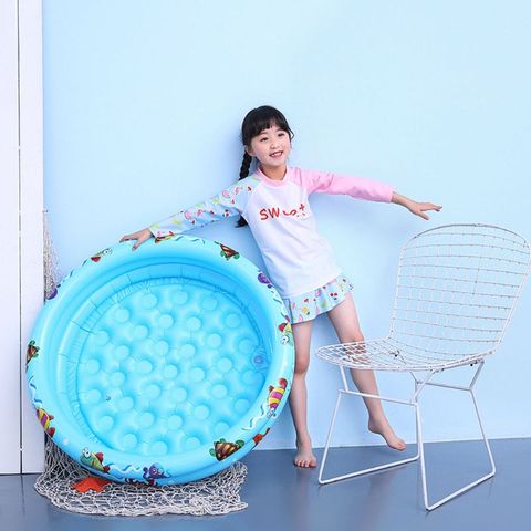 A young girl smiling, standing beside a small, blue inflatable pool with cartoon designs. She's wearing a colorful long-sleeve shirt and shorts, holding a white wire chair in a blue room.