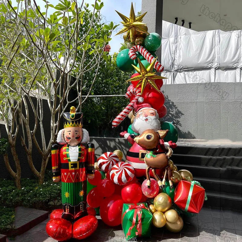 Festive outdoor scene with a nutcracker, balloons shaped like Santa, reindeer, candy canes, and gift boxes. Greenery frames the colorful Christmas decorations.