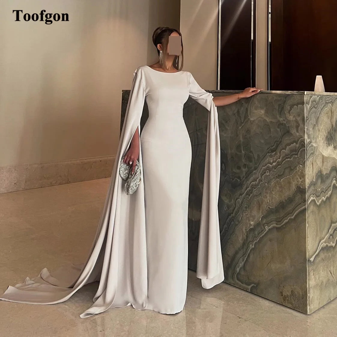 Elegant woman in a flowing white gown with cape sleeves, standing by a marble-textured wall, holding a sparkling clutch. The room has a minimalist and refined decor.