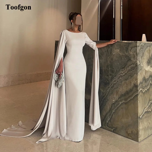 Elegant woman in a flowing white gown with cape sleeves, standing by a marble-textured wall, holding a sparkling clutch. The room has a minimalist and refined decor.