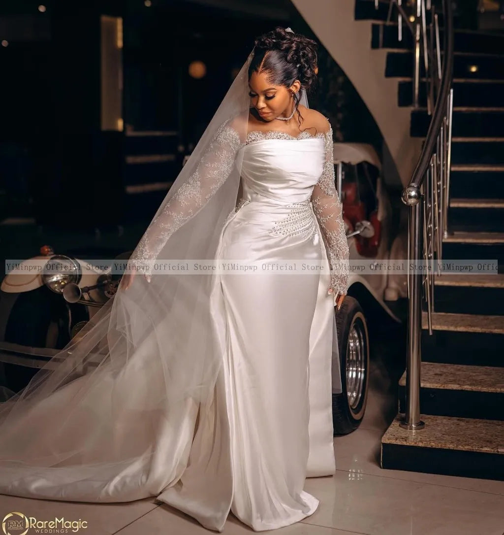 Bride in an elegant white satin gown with lace sleeves and a long veil stands near a staircase. A vintage car is partially visible in the background.