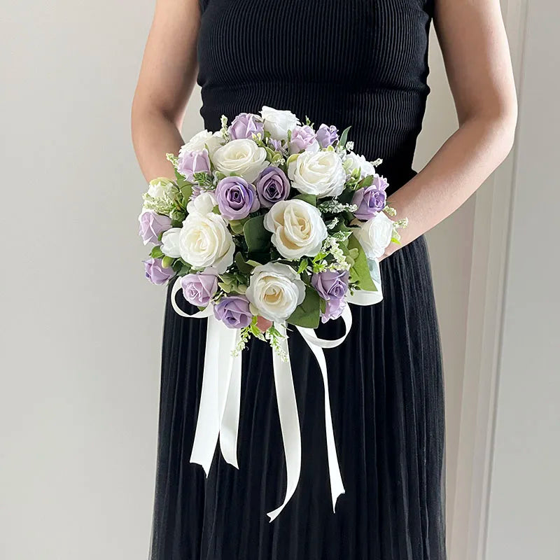 A woman in a black dress holds a bouquet of white and lavender roses, accented with green foliage and white ribbons, standing against a neutral background.