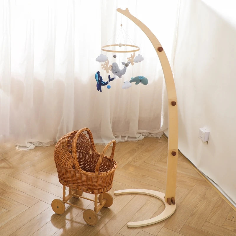 A wicker baby carriage with wooden wheels sits on a wooden floor next to a wooden stand holding a whimsical ocean-themed mobile. Sheer curtains softly filter the light.