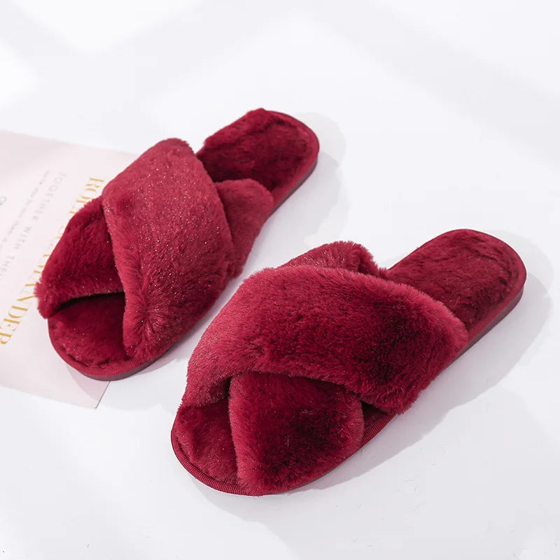 Plush burgundy slippers with crisscross straps on a white surface, offering a cozy and luxurious appearance. A partial view of text is visible on a paper underneath one slipper.