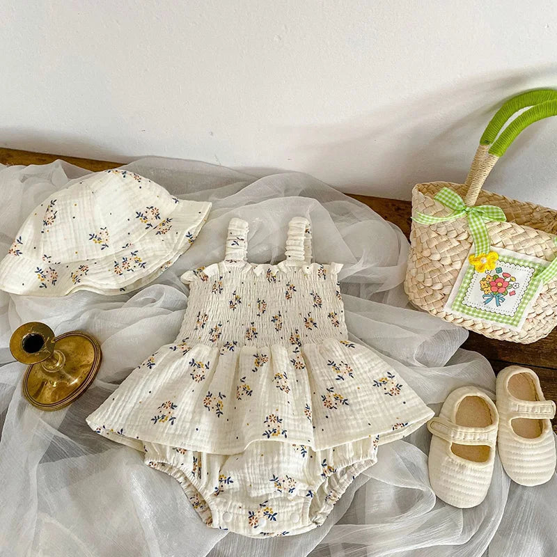 Baby outfit set with a floral-patterned romper, matching hat, cream shoes, and a woven bag with a green ribbon, set on soft fabric alongside a small brass candle holder.