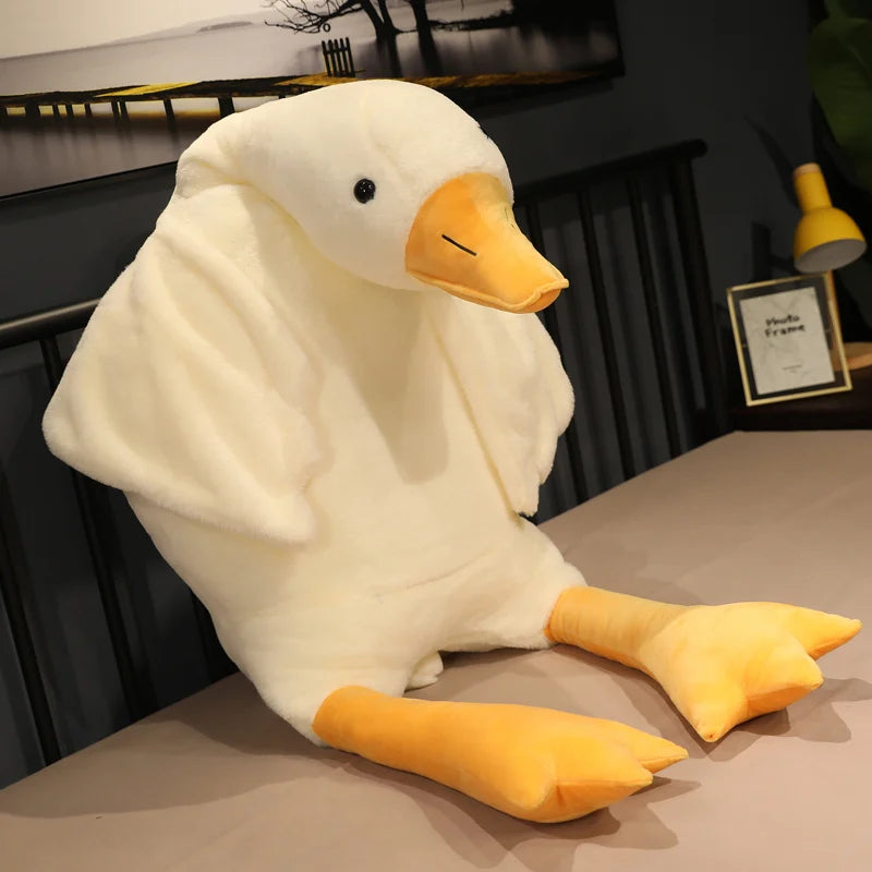 A large, plush white duck with orange feet and beak sits on a bed. The room has a dark interior with a framed picture and a lamp in the background.