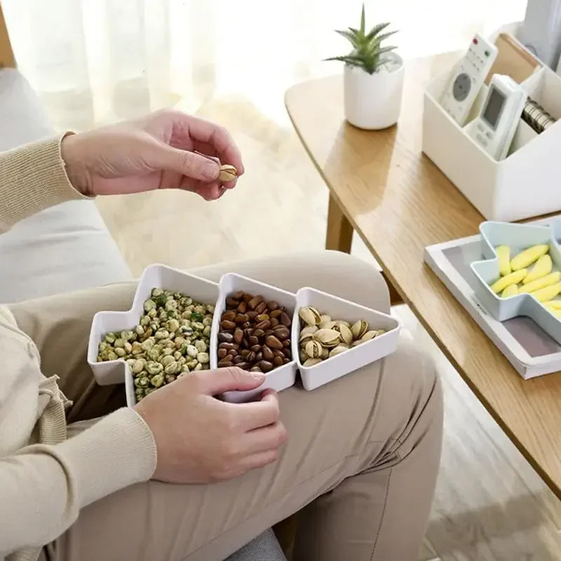 Person sitting with a divided tray holding wasabi peas, peanuts, and pistachios on their lap. A potted plant and remote controls are on a wooden table nearby.