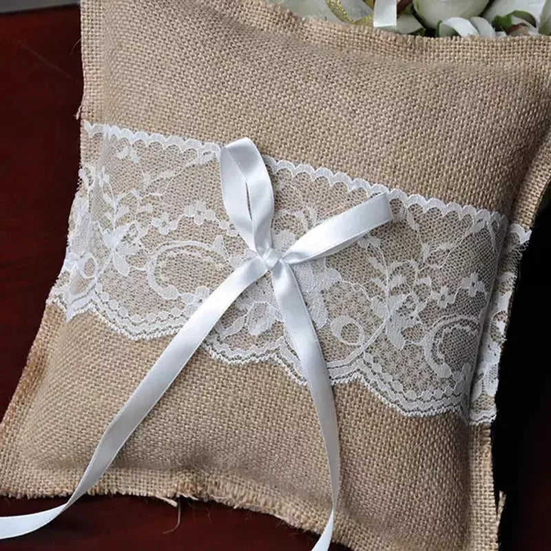 Burlap wedding ring pillow features a white lace band and satin ribbon tied in a bow. The rustic design rests on a dark wooden surface, adorned with scattered flowers.