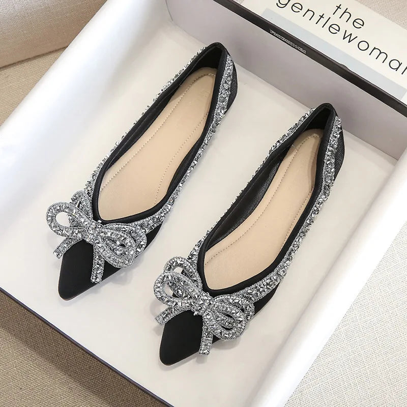 Elegant black pointed-toe flats adorned with sparkling rhinestone bows and trim, displayed in a white box.