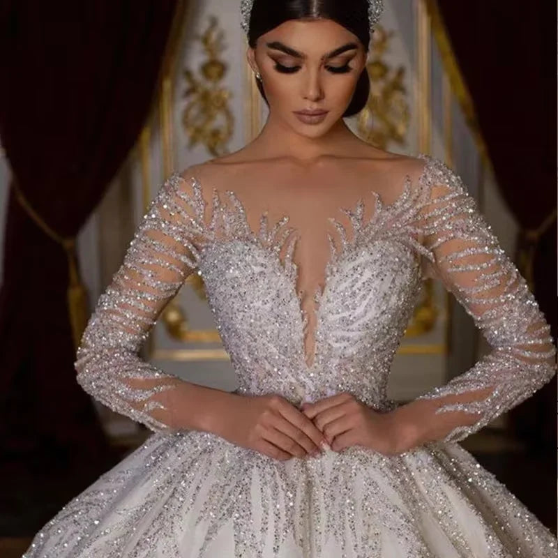 A woman in an elegant gown adorned with intricate silver beadwork stands poised in a lavish setting with opulent curtains and golden decor, her hands gently clasped.
