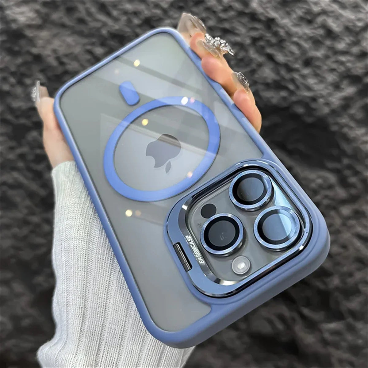 A hand holds a smartphone in a blue protective case with a visible camera array and a circular MagSafe design on the back. The person wears a sweater with long sleeves.