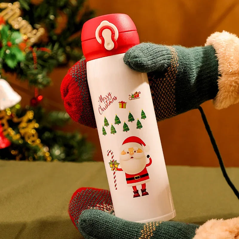 Gloved hands holding a festive thermos with Santa Claus and Christmas tree designs. A decorated tree is blurred in the background, giving a warm holiday feel.