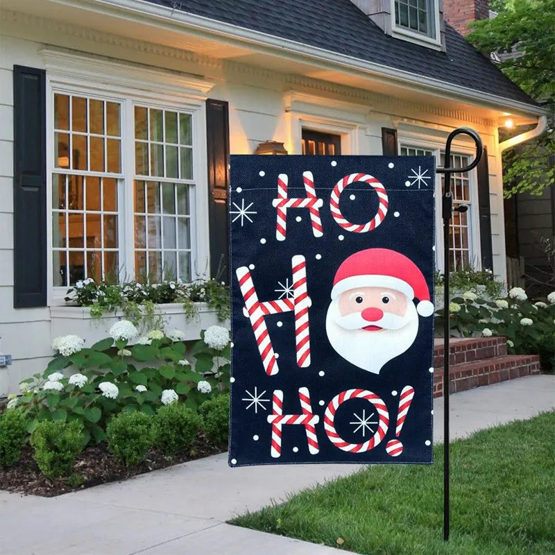 Festive garden flag with "Ho Ho Ho!" in candy cane letters and a Santa face, set in front of a white house with black shutters and a flowerbed.