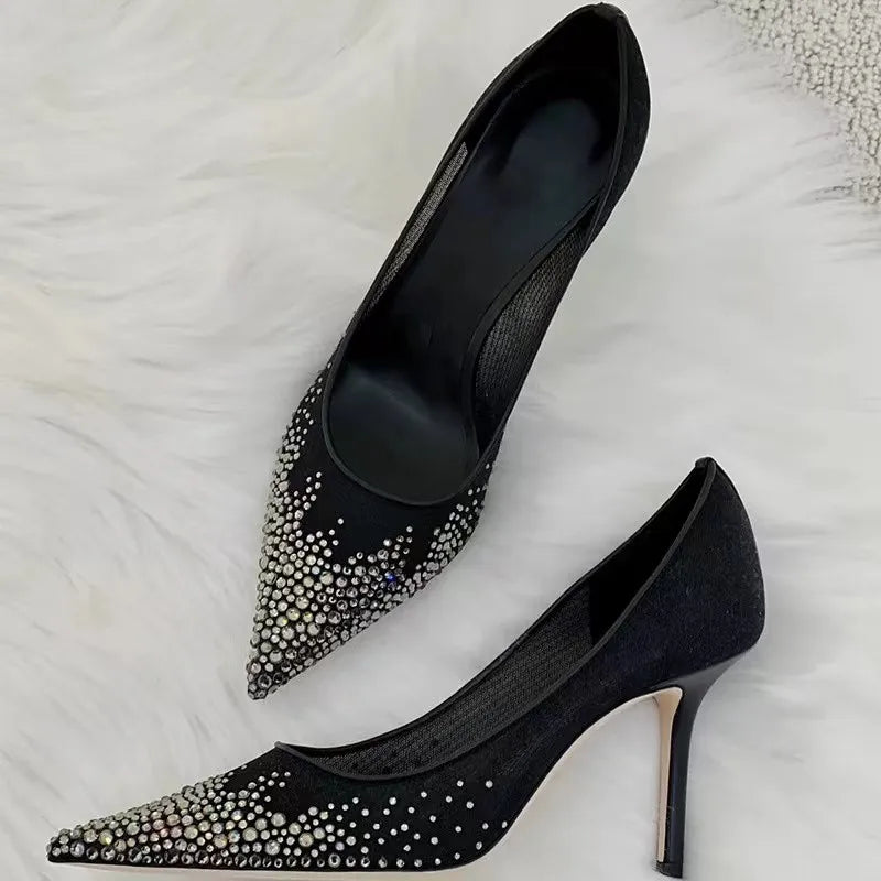 Black stiletto heels with pointed toes adorned with silver rhinestones, displayed on a white textured surface.