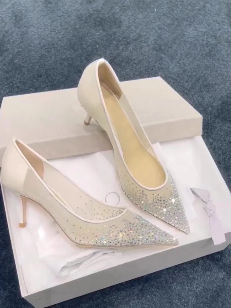 Elegant cream high heels adorned with sparkling rhinestones on a pointed toe, displayed in an open shoe box against a soft, textured gray carpet background.