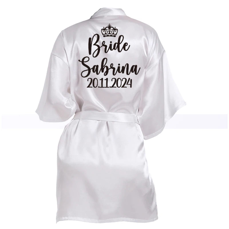 White satin robe with "Bride Sabrina 20.11.2024" written on the back in black script, featuring a crown design above the text.