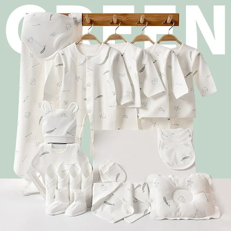 Baby clothing set on a wooden rack, featuring white garments with subtle prints, including tops, pants, bibs, mittens, a hat, and a pillow. A soft green background.