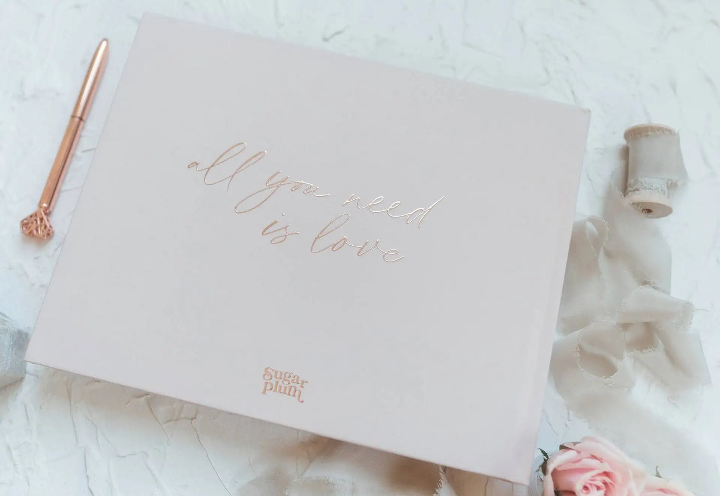 Elegant box with "all you need is love" embossed on it. Beside it, a rose gold pen, spool of ribbon, and a pink rose on a textured white surface.