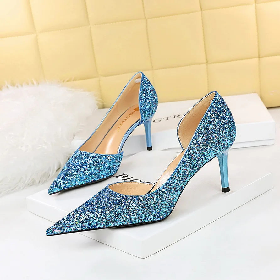 Blue glittery high heels with pointed toes and slim heels displayed on white boxes, set against a quilted cream backdrop and a fluffy white accent.