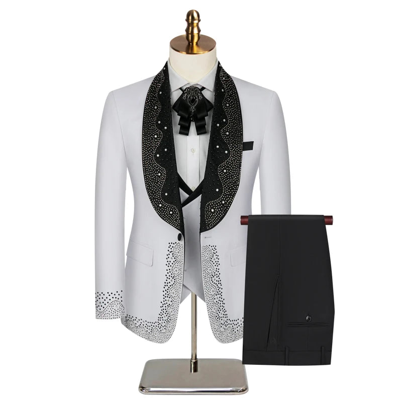 White suit with black satin lapel adorned with rhinestones, featuring a floral brooch and bolo tie. Matching black trousers beside it, displayed on a mannequin with gold head.
