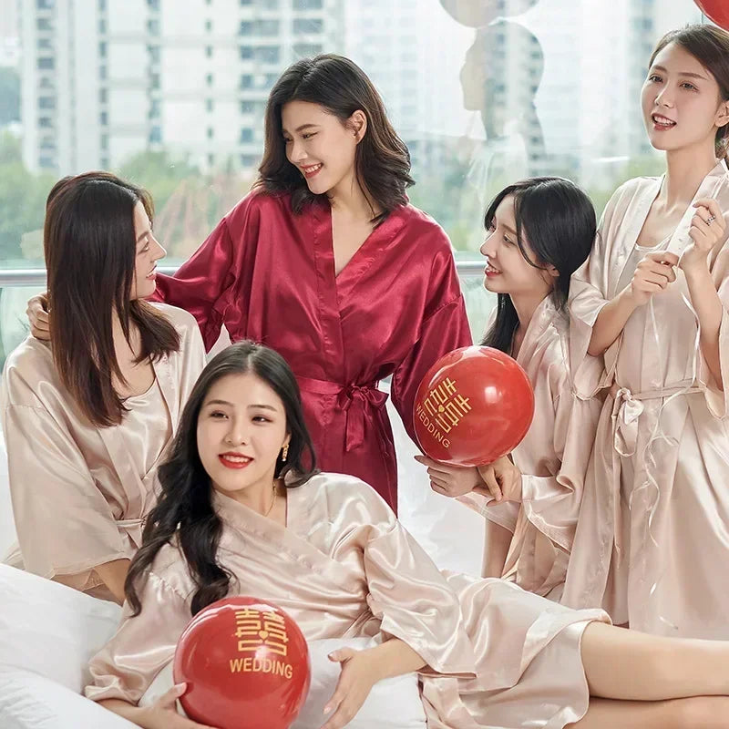 Five women in satin robes, one in red and others in beige, smile together indoors holding red balloons with “wedding” written on them. Multistory buildings are visible outside.