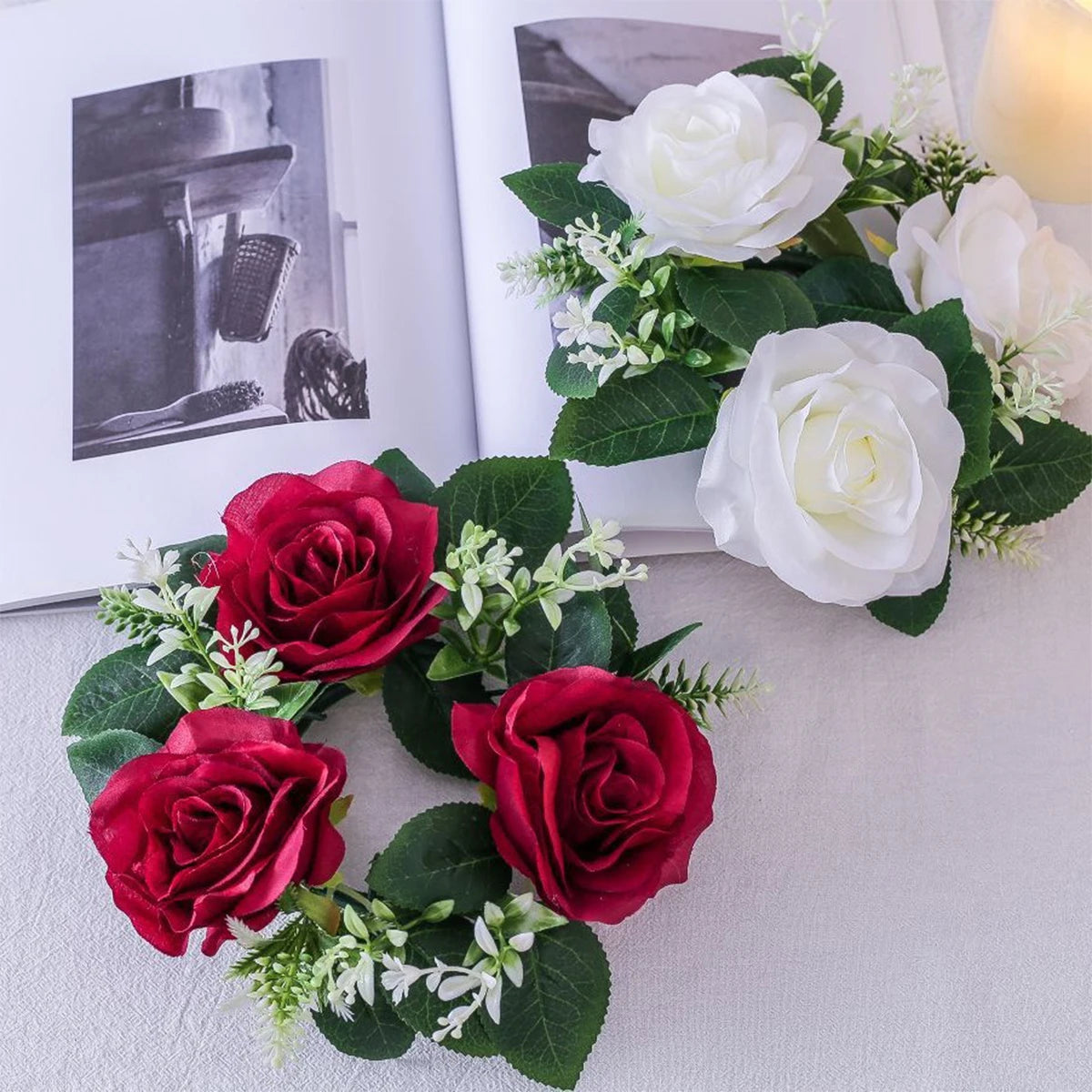 Artificial red and white roses with green leaves and small white flowers are arranged on an open book with monochrome images, set on a light-colored table.
