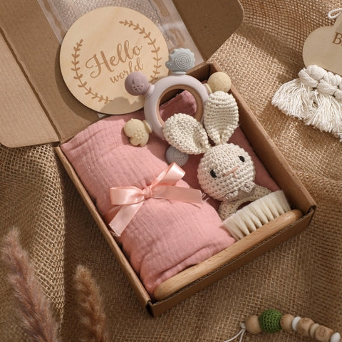A gift box contains a pink swaddle with a bow, a crocheted rabbit toy, a baby hairbrush, teething ring, and a round "Hello World" sign. The box is set on a textured fabric.