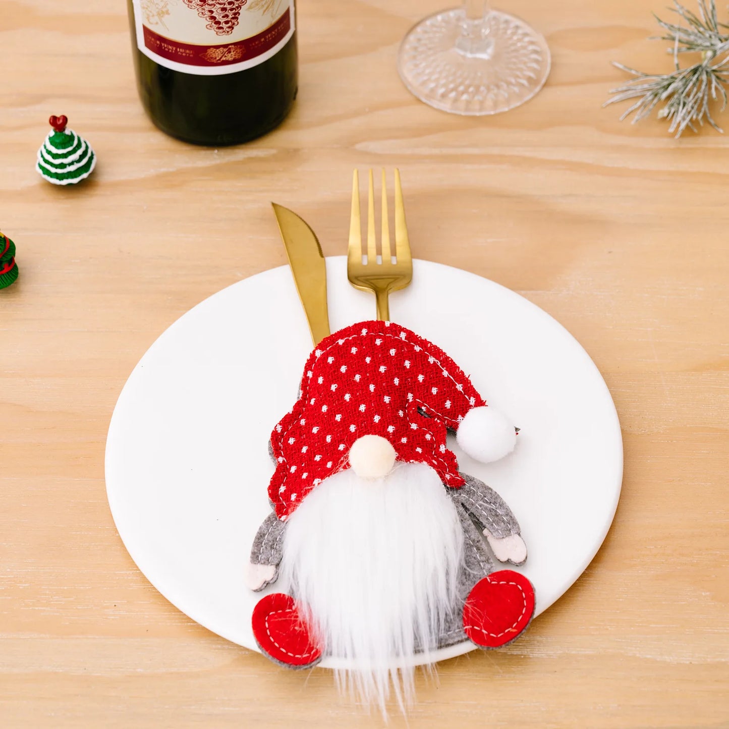 Festive table setting with a gnome figurine on a plate, gold fork and knife, bottle of wine, and mini Christmas trees on a wooden table.