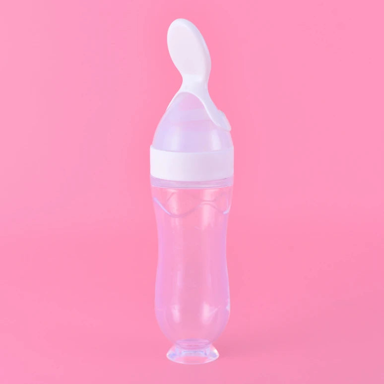 Transparent baby bottle with built-in spoon attached to a white lid, set against a solid pink background.