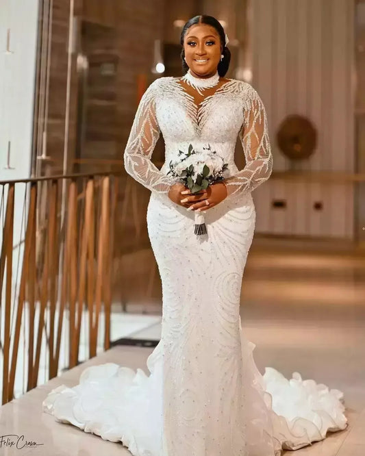 A bride in an elegant white gown with intricate beading and lace sleeves holds a bouquet. She smiles warmly, standing in a modern interior with wooden and light decor.