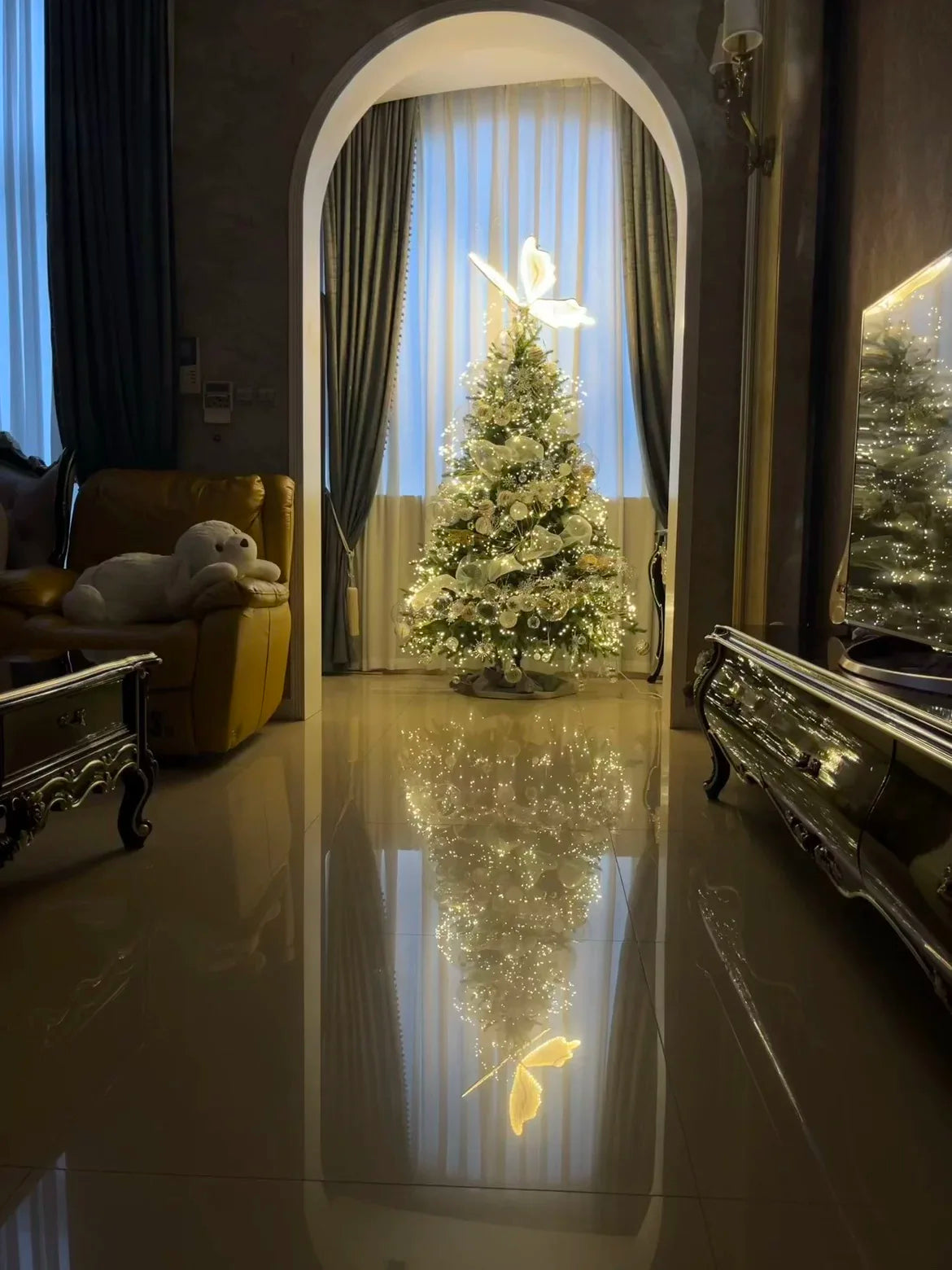 Elegant living room with a decorated Christmas tree under soft lighting. The tree's reflection shimmers on the polished floor, next to a cozy sofa with a stuffed animal.