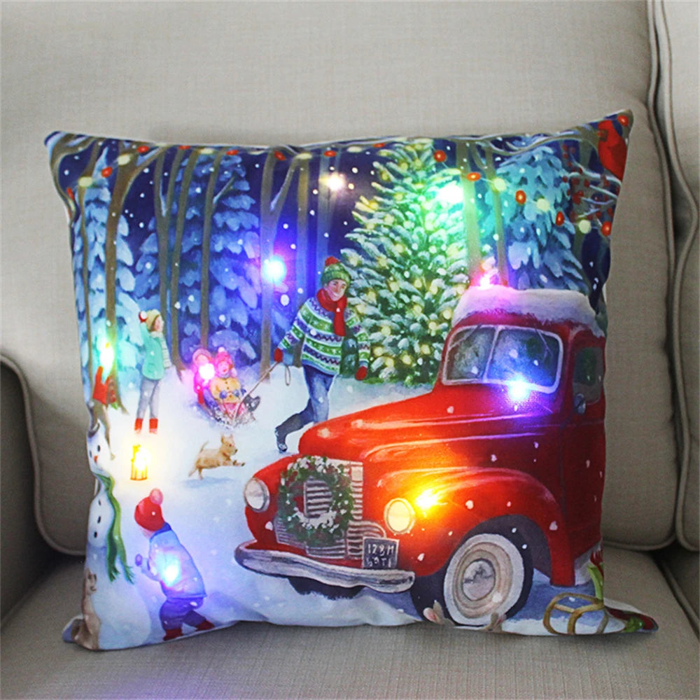 Festive cushion featuring a winter scene with people in snow, a red vintage truck adorned with a wreath, and colorful LED lights on trees, creating a cozy holiday ambiance.