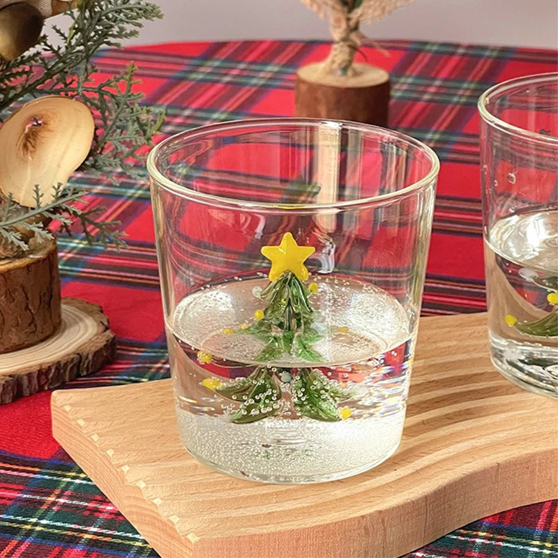 A glass with a miniature Christmas tree and yellow star inside, placed on a wooden coaster. A festive red and green plaid tablecloth is in the background with small wooden decorations.