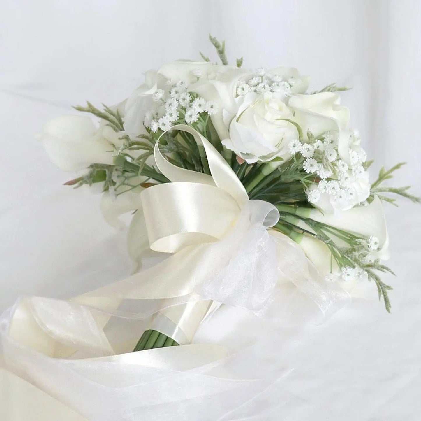 Elegant white bridal bouquet with roses, baby's breath, and greenery, tied with a satin ribbon, laying on a white fabric background.
