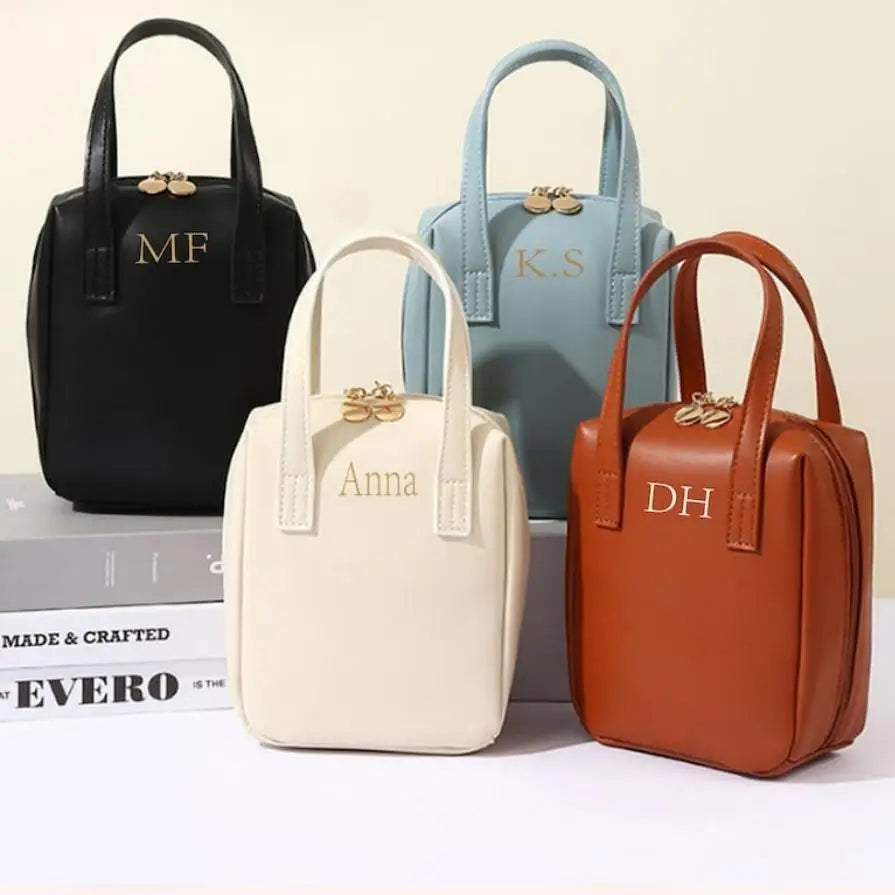 Four small handbags with gold initials: a black bag labeled "MF", light blue "K.S", cream "Anna", and brown "DH", each featuring a top handle and gold clasp.