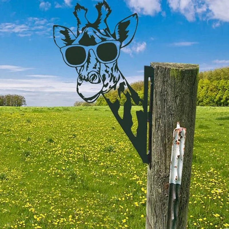 Metal cutout of a giraffe wearing sunglasses attached to a wooden post, set against a green field with yellow flowers under a blue sky with scattered clouds.