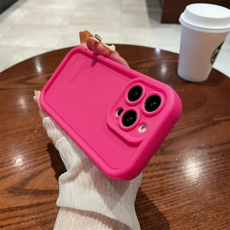 Hand holding a pink smartphone with a protective case and three camera lenses. In the background, a wooden table and a to-go coffee cup are visible.