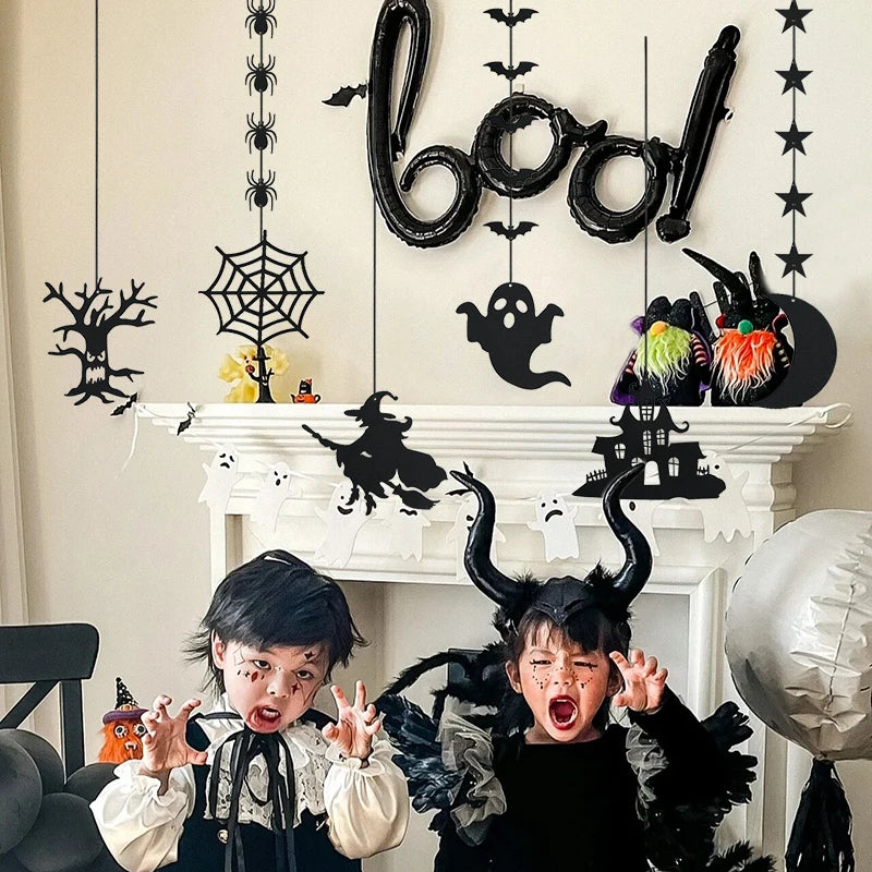 Two children in Halloween costumes pose with playful expressions in front of a decorated white mantel. The backdrop features hanging Halloween decorations, including a "boo!" sign, spiderwebs, ghosts, and a witch silhouette. The scene is festive with black and white accents, emphasizing a spooky theme.