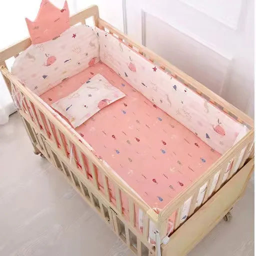 Wooden baby crib with pink and white bedding featuring whimsical designs. A pillow and crown-shaped cushion are included. The bed is placed in a bright room near white walls.