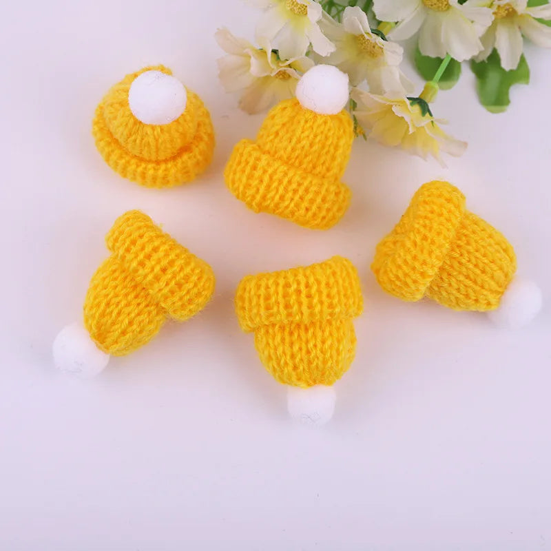 Five small knitted yellow hats with white pom-poms arranged in a circle on a light background, with white flowers partially visible in the background.