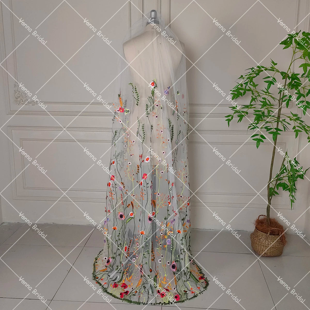 Elegant sheer bridal veil on mannequin, adorned with colorful floral embroidery. A potted plant sits nearby on a tiled floor against a paneled wall.