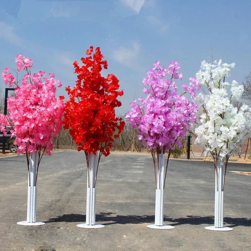 Four artificial cherry blossom trees in pink, red, purple, and white stand in a row on a paved outdoor path against a clear blue sky.