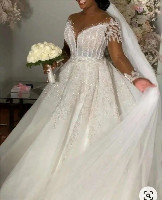 Bride in a sparkling off-shoulder gown holds a bouquet of white roses. The dress features intricate beading and long lace sleeves, with a flowing veil cascading behind.