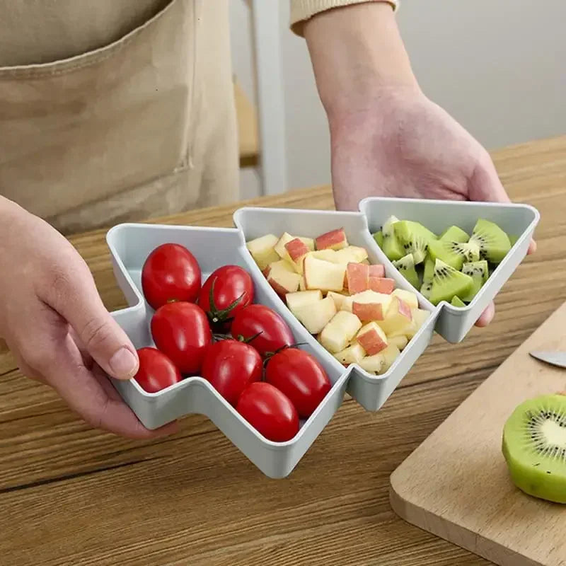 A person holds a triangular tray with compartments containing cherry tomatoes, diced apples, and kiwi slices. A cutting board with a halved kiwi is on the wooden table.