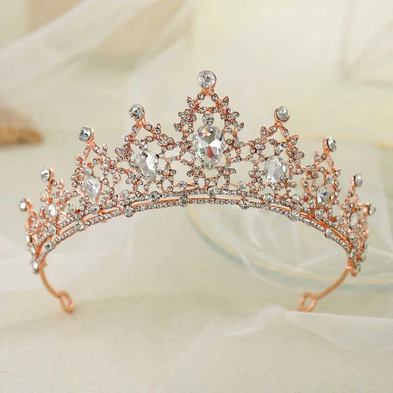 Rose gold tiara adorned with large teardrop and round crystals, set against a soft, sheer fabric background. The intricate design creates a regal and elegant appearance.