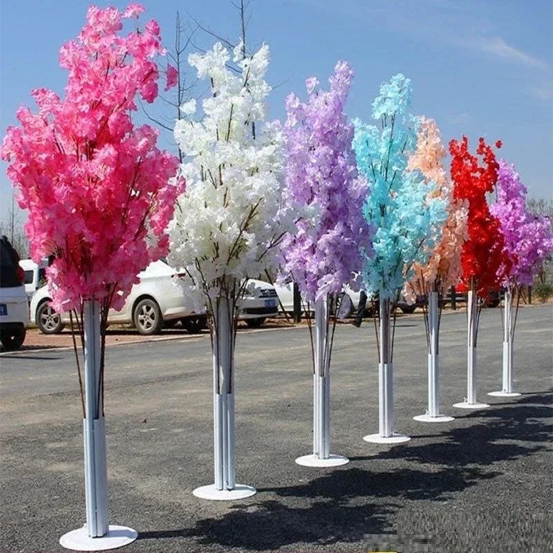 A row of artificial trees in bright colors: pink, white, purple, blue, orange, and red, stand on a paved outdoor area near parked cars under a clear blue sky.